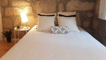 Un dormitorio con pared de piedra, una cama con ropa blanca y almohadas negras, una lámpara de noche y una planta pequeña.