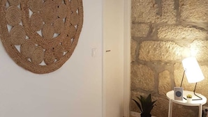 Una habitación con pared de piedra, una puerta blanca, un tapiz redondo tejido y una pequeña mesa con una lámpara y una planta.