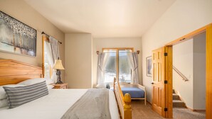 Ein Schlafzimmer mit Bett, Nachttisch, Lampe, Fenster mit Vorhängen und einer Tür zu einem anderen Raum.