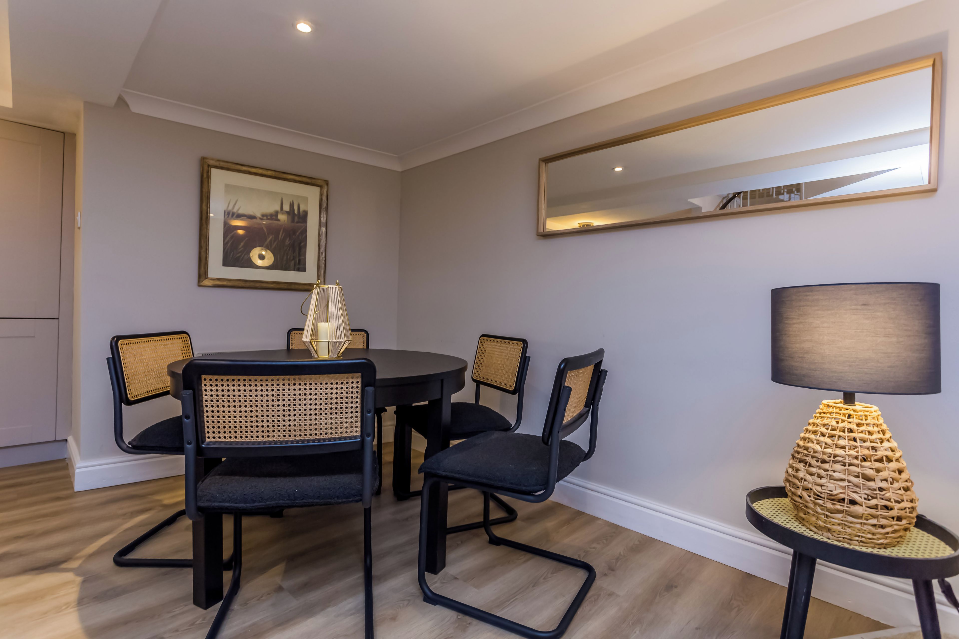 Uma sala de jantar com uma mesa redonda, quatro cadeiras, um quadro emoldurado, um espelho e um abajur.