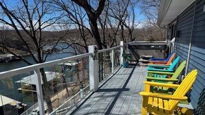 A wooden deck with a railing overlooking a body of water, colorful Adirondack chairs, and a view of a lake and trees.