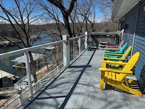 A wooden deck with a railing overlooking a body of water, colorful Adirondack chairs, and a view of a lake and trees.