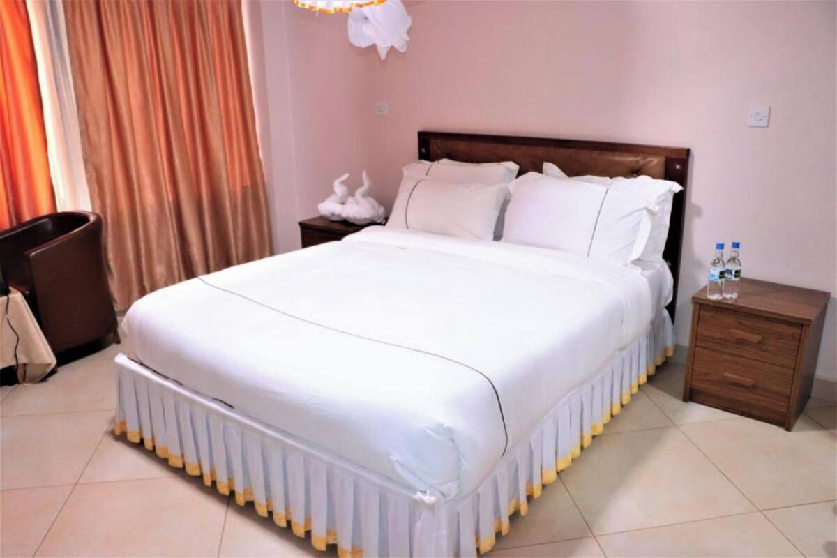 A hotel room with a large bed, a wooden headboard, a nightstand with a lamp, and a small table with water bottles.