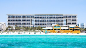 Un gran hotel con varios pisos y balcones, una playa con sombrillas y sillas de descanso, y un mar azul y transparente.