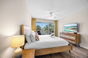 Een slaapkamer met een bed, een televisie, een plafondventilator, een raam met gordijnen en uitzicht op de omgeving.