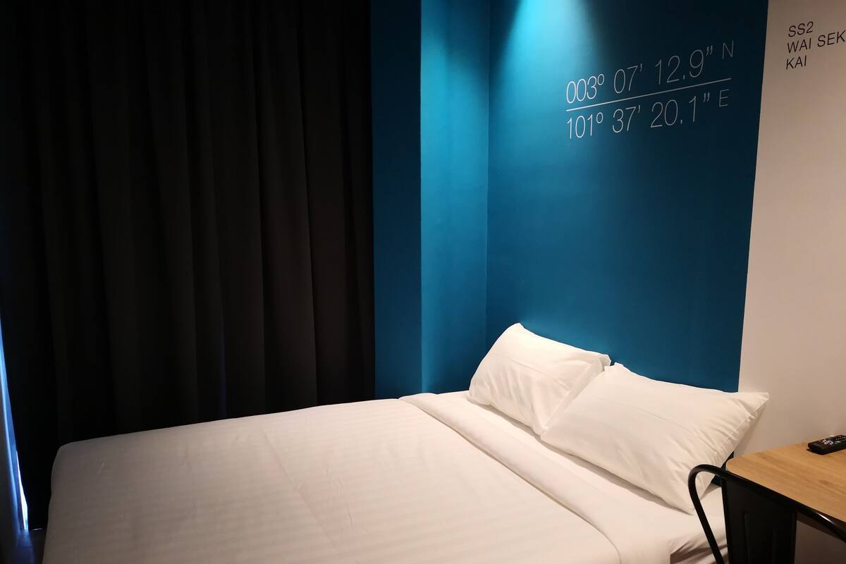 A hotel room with a bed, a wooden nightstand, and a wall displaying coordinates.
