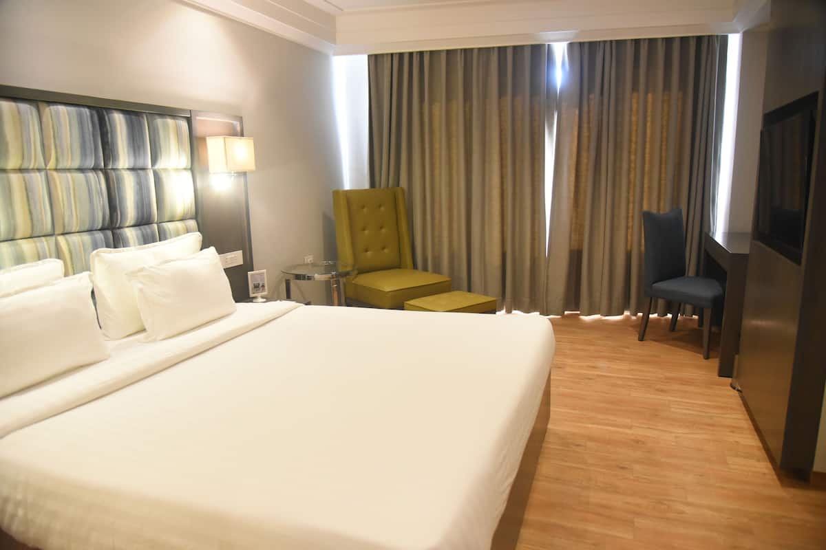Ein Hotelzimmer mit einem großen Bett, einem Fernseher, einem Schreibtisch mit Lampe, einem Sessel und einem Fenster mit Vorhängen.