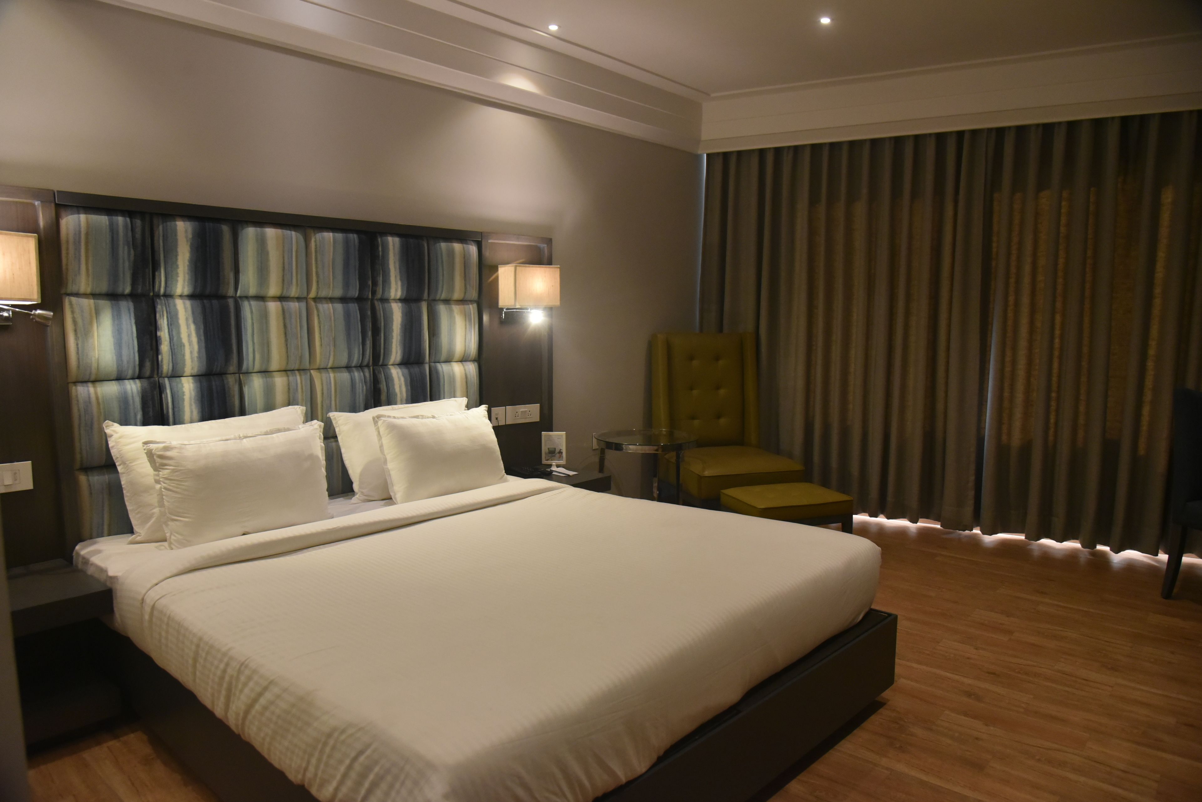 Une chambre d’hôtel avec un grand lit, une tête de lit à rayures verticales, une lampe de chevet, une chaise et une fenêtre avec des rideaux.