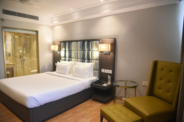 Una habitación de hotel con una cama grande, una mesita de noche con lámpara, una mesa de vidrio y un sillón verde.