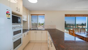 A modern kitchen with white cabinets, a built-in microwave, and a stove. There is a large window with blinds, and a dining area with a table and chairs. - Seacrest Unit 4 Moffat Beach QLD (MOFFAT BEACH)