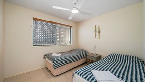 A bedroom with two beds, a ceiling fan, a window with blinds, and a bedside table with a lamp. - Seacrest Unit 4 Moffat Beach QLD (MOFFAT BEACH)