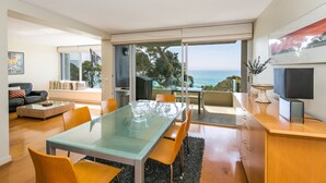 A modern living room with a glass dining table, comfortable seating, a television, and a view of the ocean. - Apartment 7 Kalimna (Lorne)