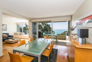 A modern living room with a glass dining table, comfortable seating, a television, and a view of the ocean. - Apartment 7 Kalimna (Lorne)