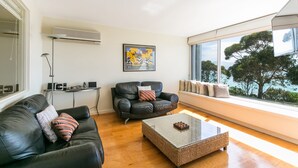 A modern living room with a black leather sofa, a glass coffee table, and a view of trees through large windows. - Apartment 7 Kalimna (Lorne)