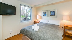 A hotel room with a bed, a flat-screen TV mounted on the wall, two framed artworks, a bedside lamp, and a window with a view of greenery. - Apartment 7 Kalimna (Lorne)