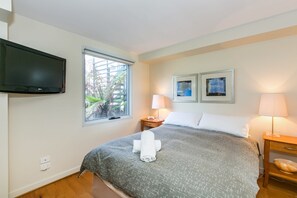 A hotel room with a bed, a flat-screen TV mounted on the wall, two framed artworks, a bedside lamp, and a window with a view of greenery. - Apartment 7 Kalimna (Lorne)