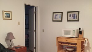 Ett vardagsrum med trägolv, en grön soffa, ett träbord med en lampa, ett träskåp med köksartiklar och inramade tavlor på väggen.