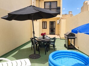 Un patio con una mesa redonda, sillas, un paraguas negro, una parrilla y una piscina inflable azul.