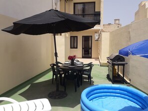 Un espace patio comprenant une table ronde, des chaises, un parasol noir, une piscine gonflable bleue et un barbecue.