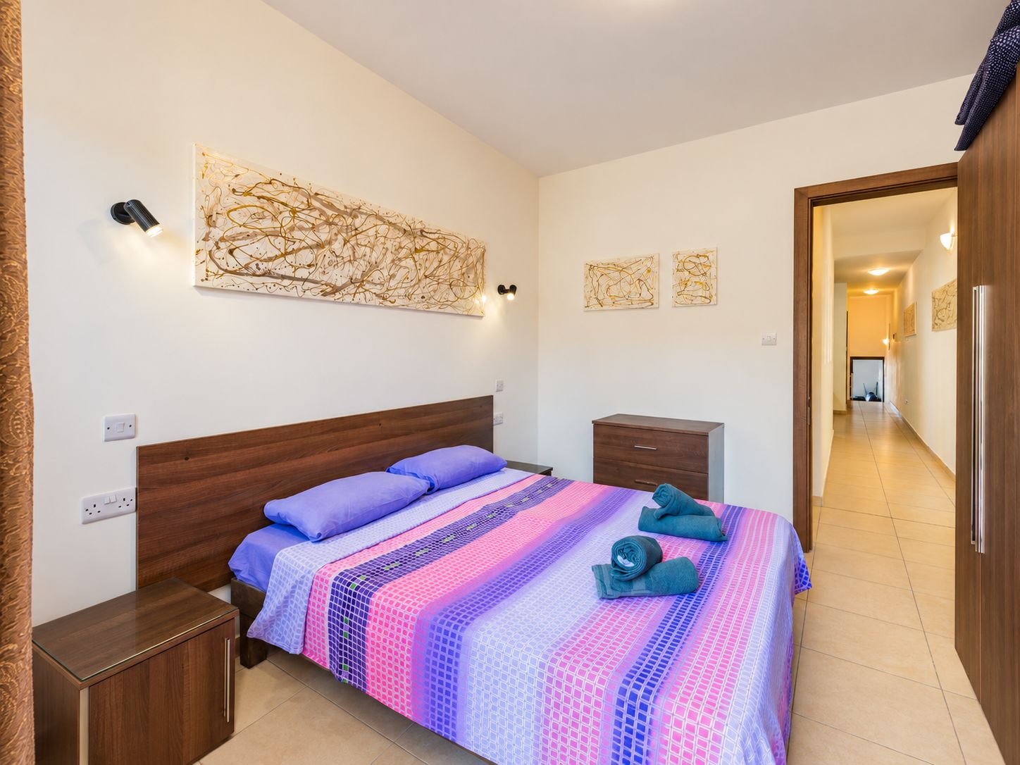 Ein Schlafzimmer mit einem hölzernen Kopfteil, einem Bett mit einem gemusterten Bettbezug, einem Nachttisch, einer Kommode und Wandbildern.