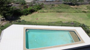 Une piscine rectangulaire, bordée d’une terrasse carrelée, entourée d’une clôture et d’une zone herbeuse avec des arbres.