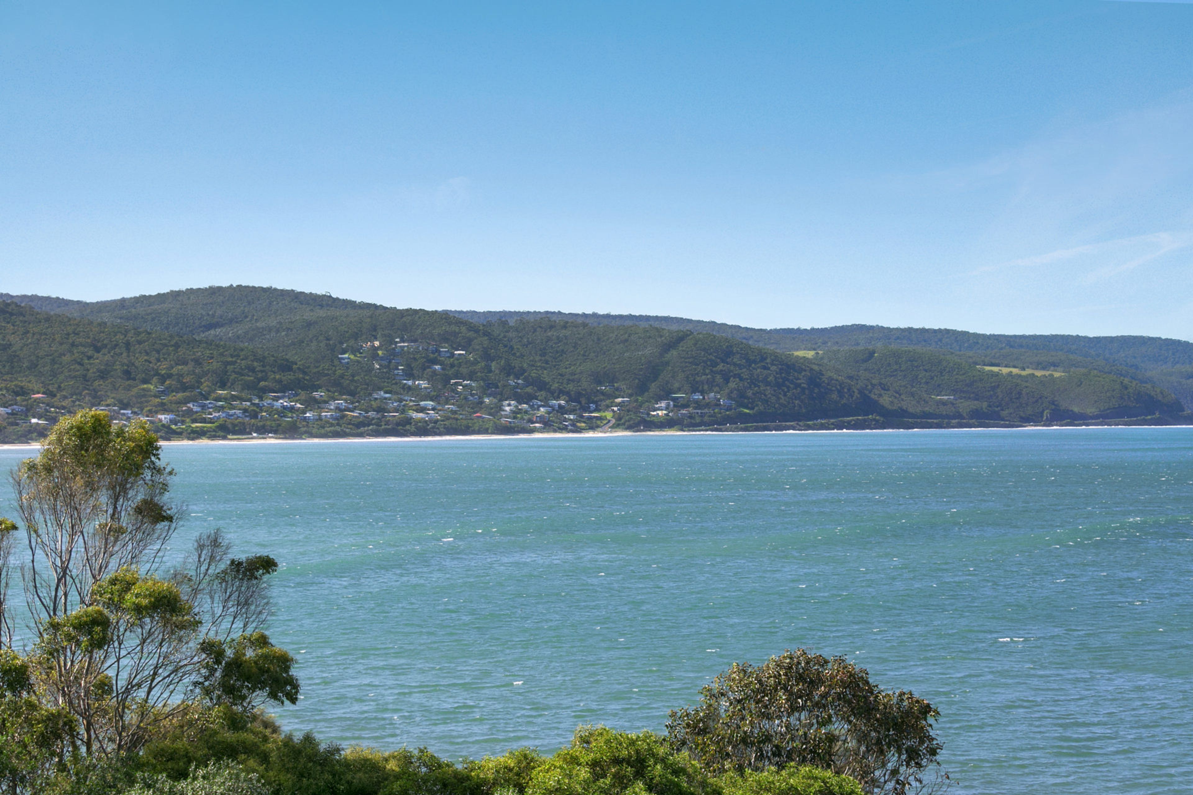 A coastal landscape with a clear view of the sea, a forested hill, and a distant shoreline with buildings.
