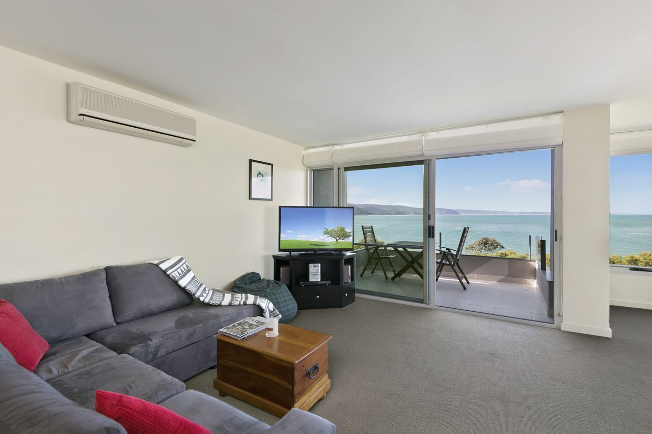A modern living room with a grey sofa, a coffee table, and a flat-screen TV. There is a sliding glass door leading to a balcony with a view of the sea.