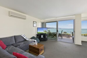 Un salon moderne avec un canapé gris, une table basse et un téléviseur à écran plat. Une porte-fenêtre coulissante donne accès à un balcon avec vue sur la mer.