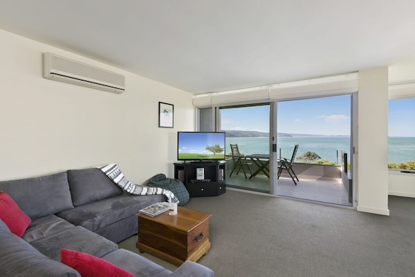 A modern living room with a grey sofa, a coffee table, and a flat-screen TV. There is a sliding glass door leading to a balcony with a view of the sea.