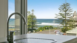 A window view of a coastal road with a clear sky, a few cars, and a large tree. - Apartment 9 Kalimna (Lorne)