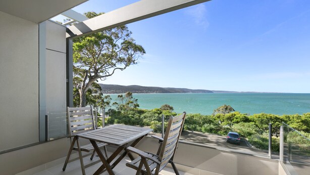 A balcony with a table and chairs, overlooking a coastal landscape with a clear view of the sea and distant hills. - Apartment 9 Kalimna (Lorne)