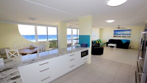 A modern kitchen with a large island, a dining area with a view of the ocean, and a living room with a sofa and a painting. - Culverston Unit 15 Kings Beach QLD (KINGS BEACH)