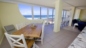 A dining area with a wooden table and white chairs, overlooking a beach with the ocean view. - Culverston Unit 15 Kings Beach QLD (KINGS BEACH)