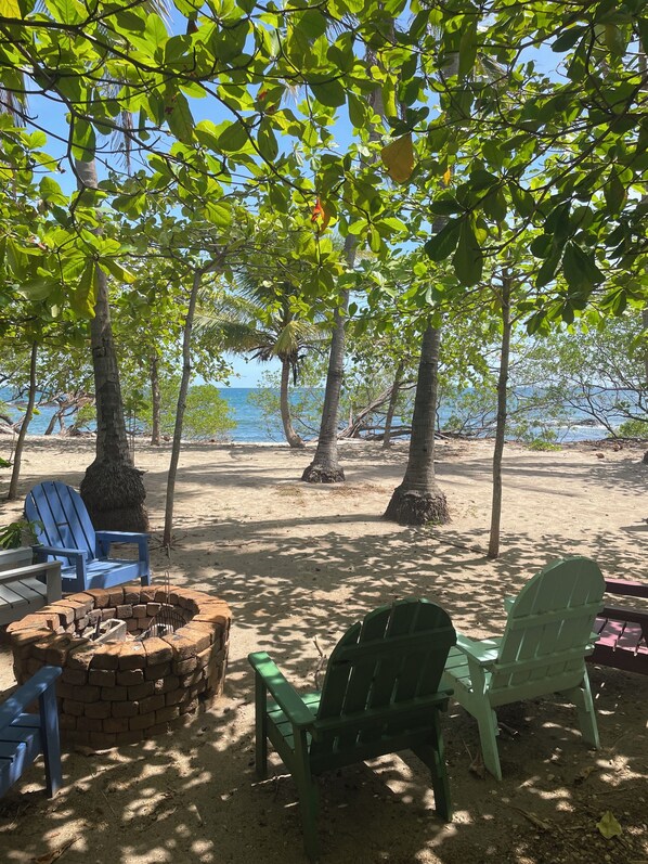 Un espace aménagé avec des sièges et un foyer, situé au bord de la plage, entouré d’arbres et offrant une vue sur la mer.
