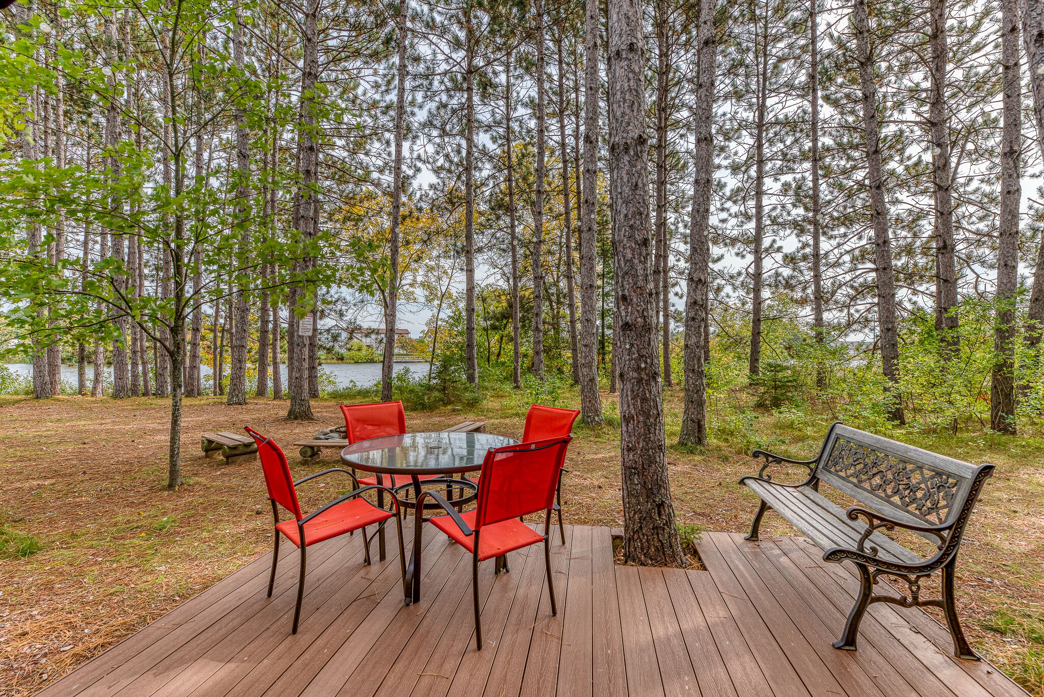 A wooden deck with a glass table and red chairs, surrounded by tall trees and a bench.