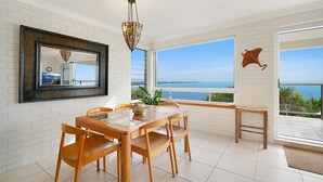 A dining area with a table and chairs, a hanging light fixture, a large window with a view of the sea, and a wall-mounted mirror.
