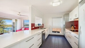 A modern kitchen with white cabinetry, a large island, and a view of the sea through a sliding door. - Moondara Unit 5, Bulcock Beach QLD (CALOUNDRA)