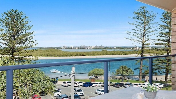 A balcony with a view of a beach, parking lot, and distant buildings. - Moondara Unit 5, Bulcock Beach QLD (CALOUNDRA)