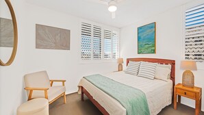 A bedroom with a bed, a chair, a bedside table, and a window with blinds. - Linden Terrace Unit 2 Kings Beach QLD (CALOUNDRA)