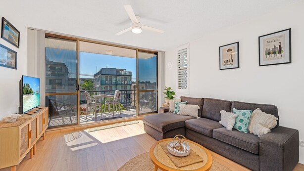 A modern living room with a grey sofa, a wooden coffee table, and a TV on a wooden cabinet. Large windows offer a view of buildings and a balcony. - Linden Terrace Unit 2 Kings Beach QLD (CALOUNDRA)