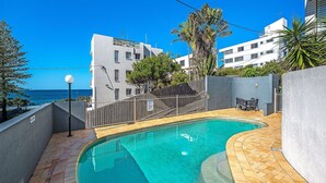 A swimming pool with a tiled deck, surrounded by a low fence, and overlooking a body of water with buildings in the background. - Linden Terrace Unit 2 Kings Beach QLD (CALOUNDRA)