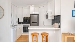 A modern kitchen with white cabinetry, a stainless steel refrigerator, a built-in oven, and a microwave. There are two wicker stools at the counter. - Linden Terrace Unit 2 Kings Beach QLD (CALOUNDRA)