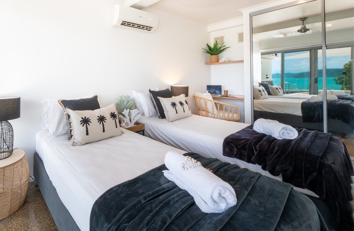 A hotel room with two beds, a desk, a chair, a large mirror, and a view of the sea.