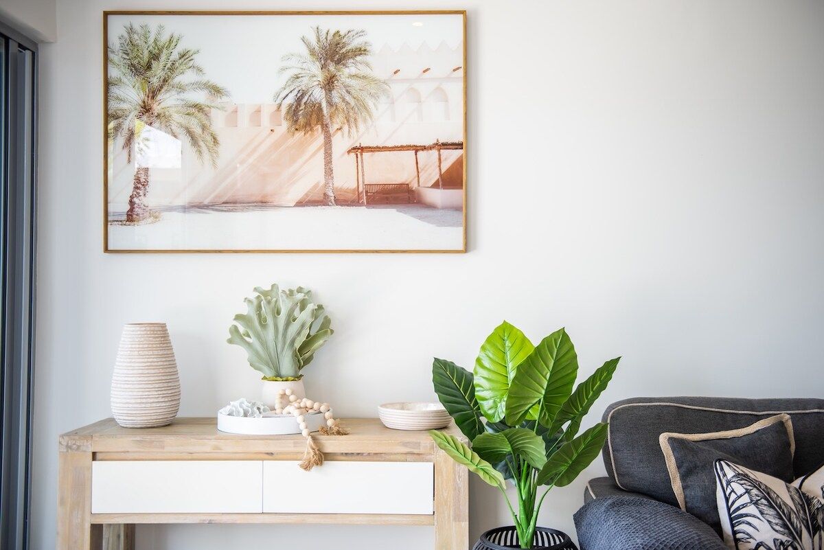 Ein Zimmer mit einem gerahmten Bild von Palmen, einem Holzkonsolentisch mit Dekoration und einer großen, grünen Pflanze.