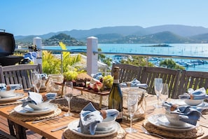 An outdoor dining area with a view of the sea and hills, equipped with tables, chairs, and a grill.