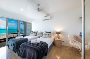 A hotel room with two beds, a desk, a chair, a lamp, and a view of the sea.