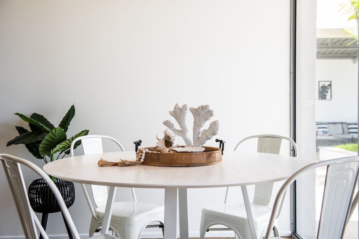 A round white table with a wooden tray and decorative item, surrounded by white chairs.