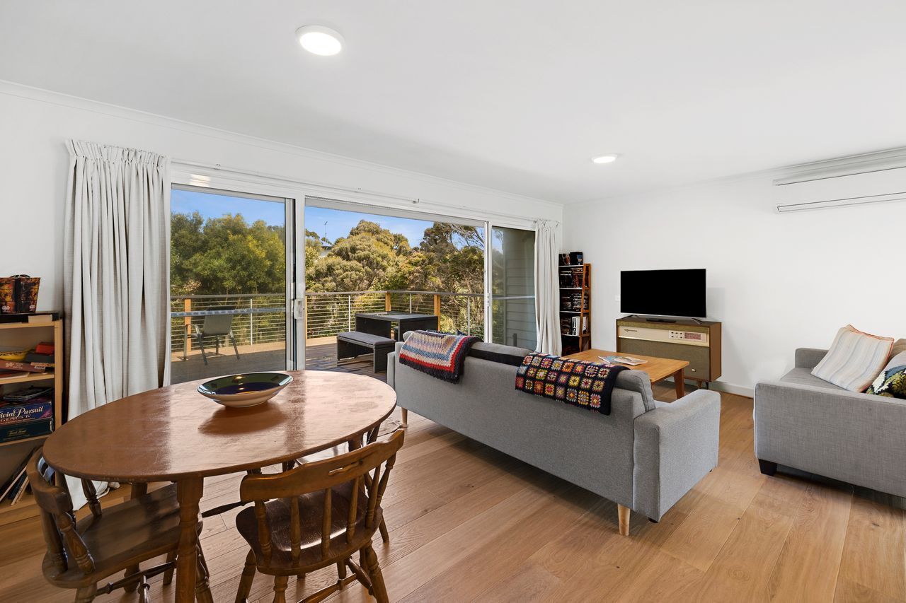 A modern living room with a sofa, a TV, a dining table, and a balcony with a view of trees.