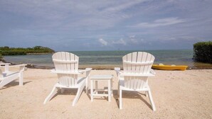 Zwei weiße Adirondack-Stühle und ein kleiner Tisch am sandigen Strand mit Blick auf das Meer und eine entfernte Insel.