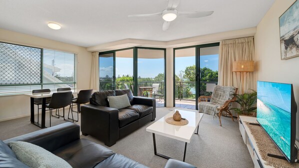 Ein modernes Wohnzimmer mit einer Ledersofa, einem weißen Couchtisch und einem Fernseher. Es gibt einen Balkon mit Tisch und Stühlen sowie ein großes Fenster mit Jalousien.